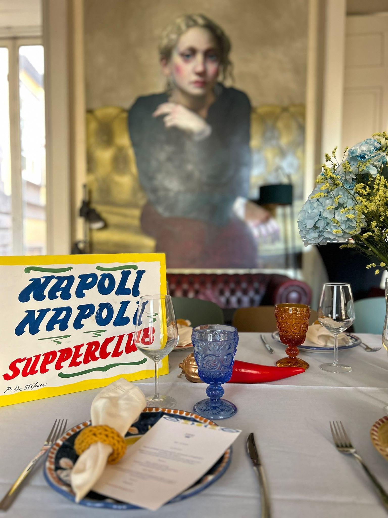 A long table laid under a painting: the hand-painted Napoli Napoli Supperclub sign, coloured glasses and a menu at every place